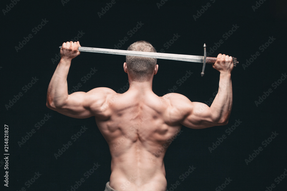 Bodybuilder man posing with a sword isolated on black background. Serious shirtless man demonstrating his mascular body. sword for head Bodybuilder man posing with a sword isolated on black background. Serious shirtless man demonstrating his mascular body. sword for head