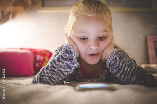 Beautiful blond toddler girl laying on her bed watching videos on a smart phone