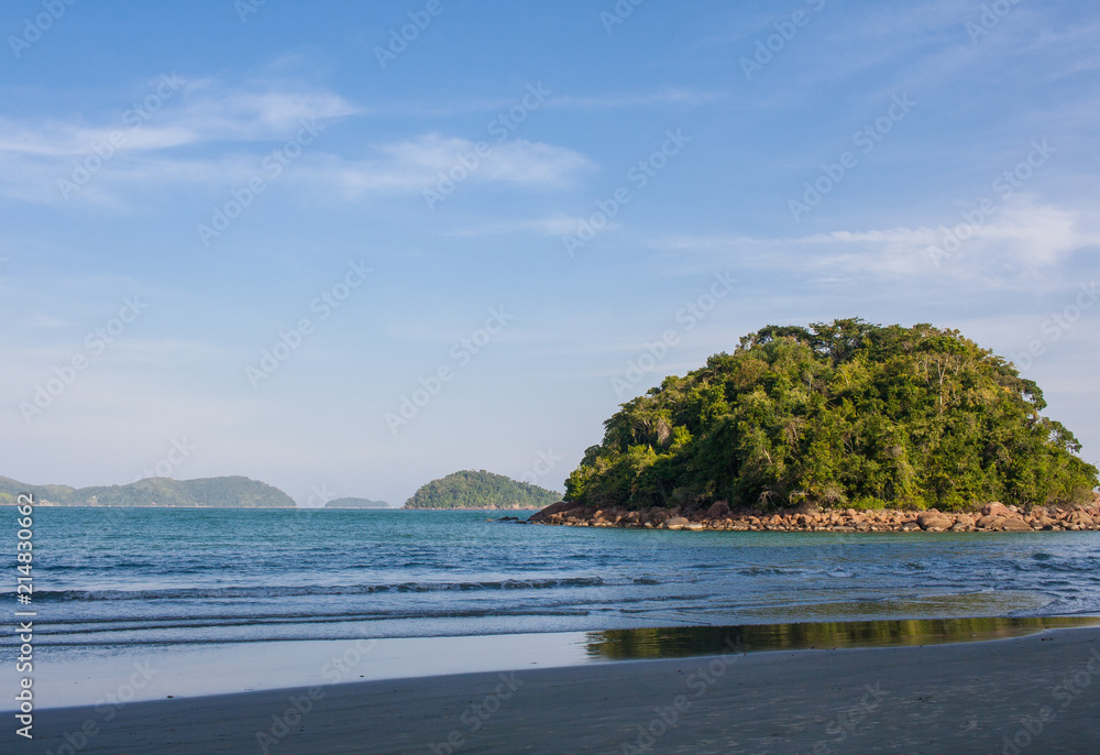 Naklejka premium Ubatumirim, Ubatuba, Brazil - Empty and calm beach, with blue and turquoise water, blue sky, an isolated island and clouds