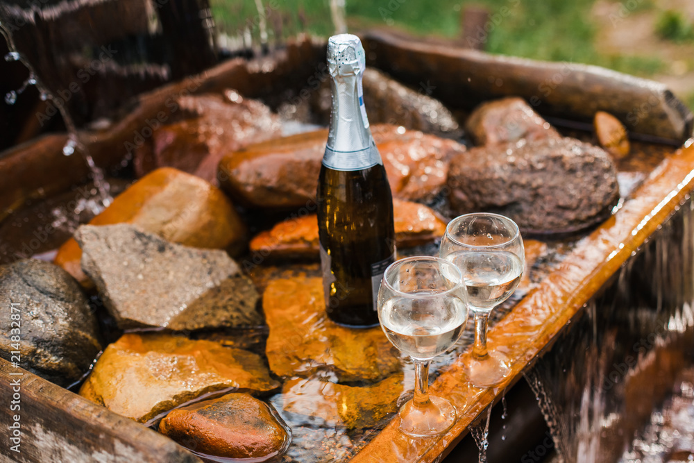 Love, romance, holiday, celebration concept. Bottle and glasses of champagne chilled by waterfall in summer forest on sunset.