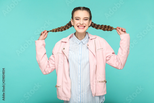 funny smilely, portrait of beautiful cute girl standing and holding her brown pigtail hairstyle in striped light blue shirt pink jacket. indoor, studio shot isolated on blue or green background. photo