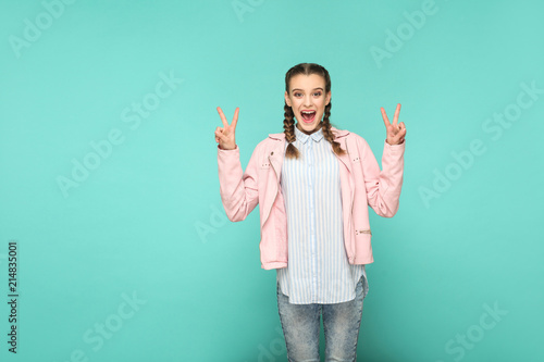 peace or victory portrait of beautiful cute girl standing with makeup and brown pigtail hairstyle in striped light blue shirt pink jacket. indoor, studio shot isolated on blue or green background.