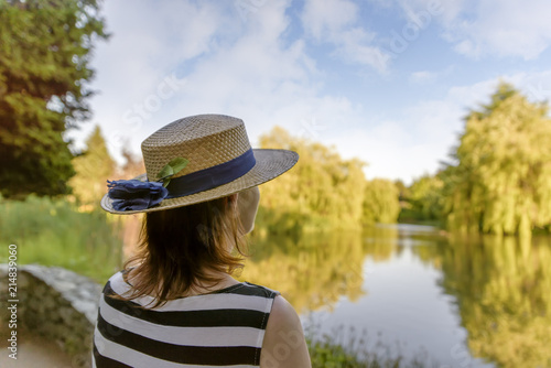 woman in a straw hat, standing in a park by the pond and admiring the nature