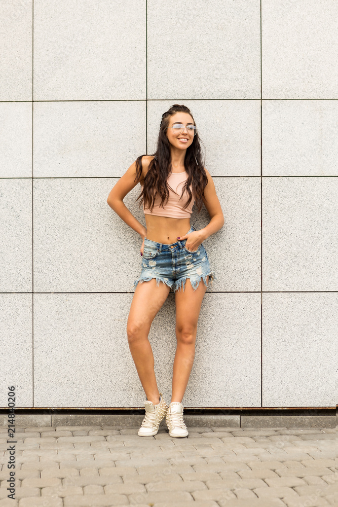 Portrait of athletic young cute woman in denim shorts, pink top and fashion  glasses smiling and have good time. Urban style, healthy girl lifestyle and  sport concept. Hipster city life. Stock Photo