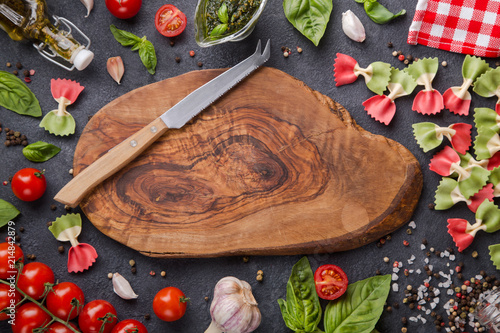 Italian flag farfalle pasta on dark background with copy space on wood cut board horizontal. Cheese knife, herry tomatoes, garlic, basil, pesto, olive oil, pepper mix, salt and red napkin photo