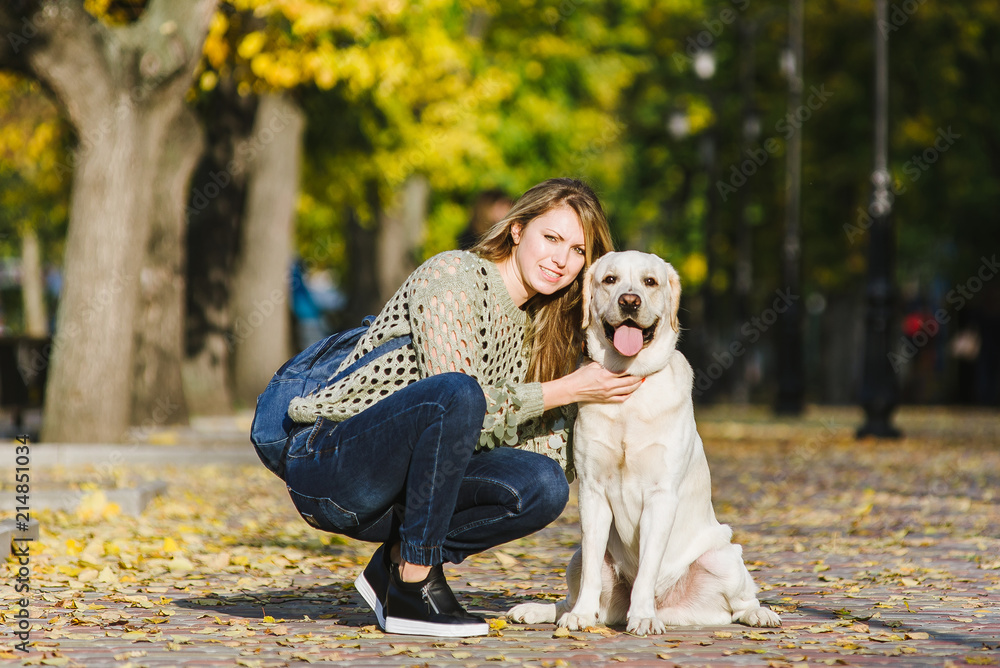 Beautiful young blonde is walking in the park with her labrador in the park in the fall. The woman crouched beside her retriever. Beautiful young blonde is walking in the park with her labrador in the park in the fall. The woman crouched beside her retriever.