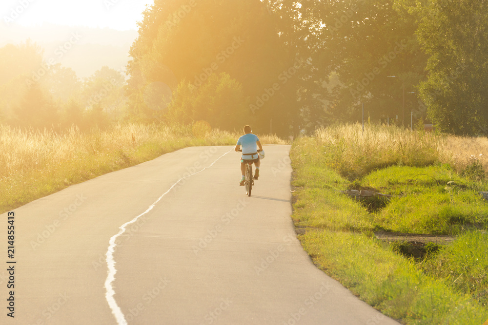 A young guy rides a bicycle on an asphalt road, leaves in the distance, the evening sunset