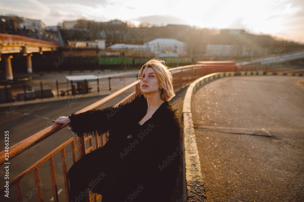 Young attractive hipster woman with colorful make-up in black fur coat posing outdoors on the road on sunset. Urban background.