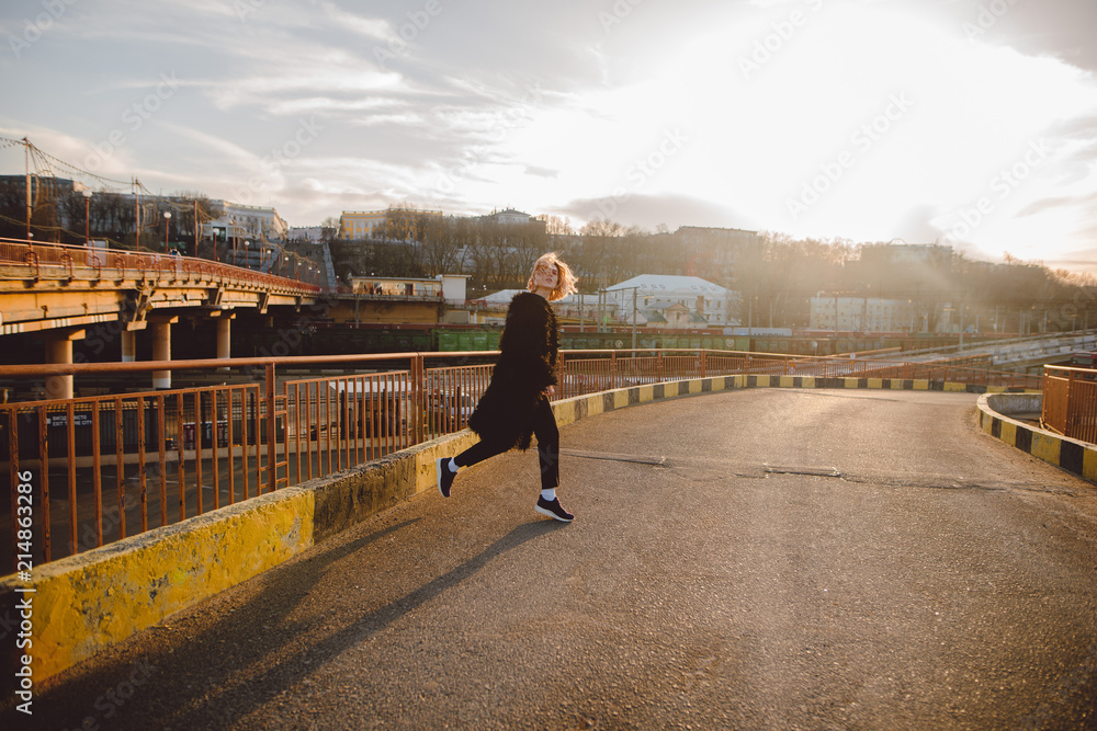 Fototapeta premium Attractive blonde hipster woman in black fur coat running outdoors on the road on sunset. Urban background. Grunge street style