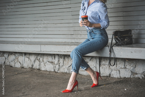 Girl in red shoes and blue jeans sitting on the banch and drinking coffee 
