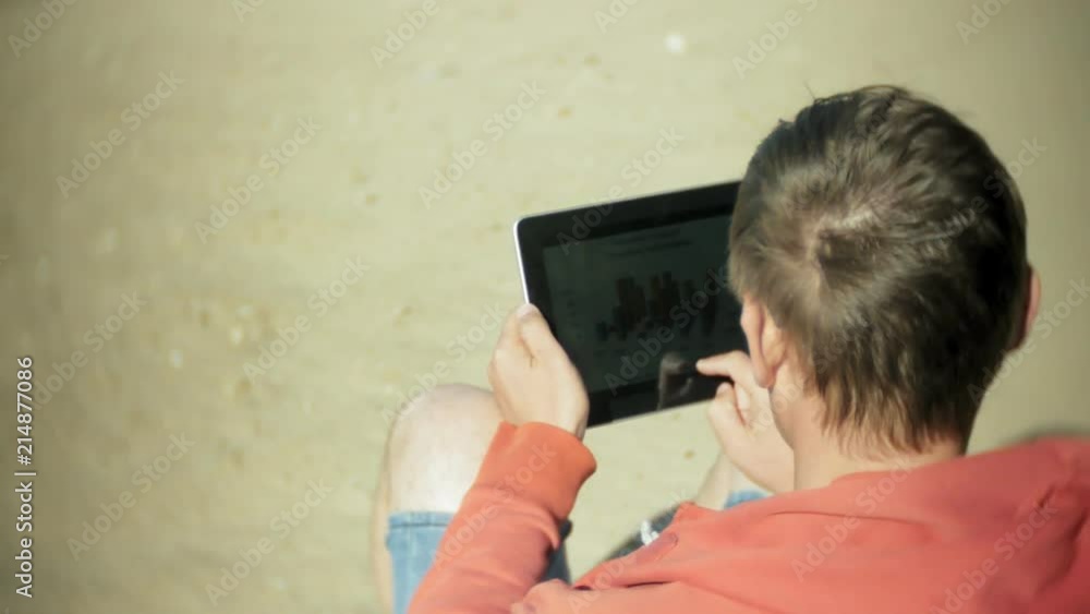 Old man sits on the beach in headphones near the boat and uses a tablet Old man sits on the beach in headphones near the boat and uses a tablet
