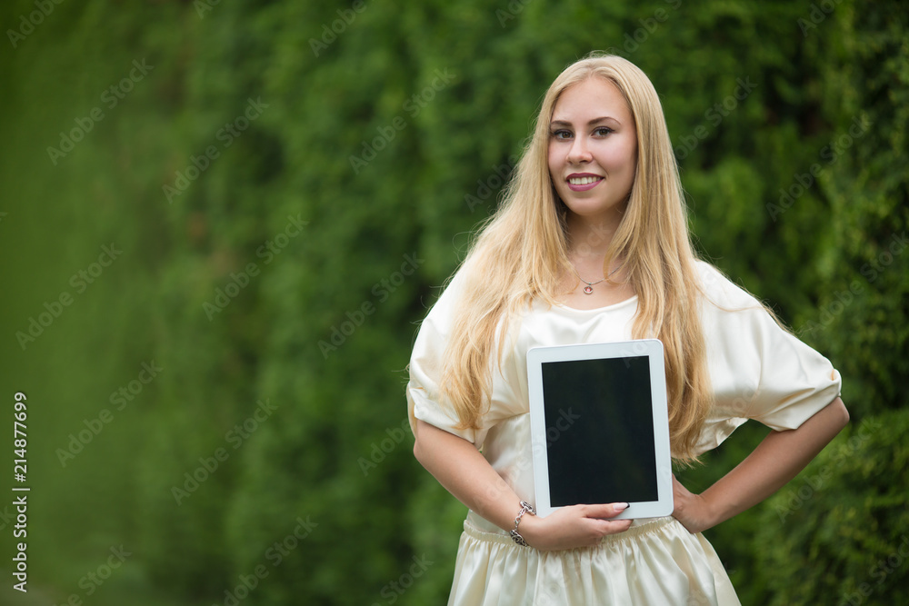 Young pretty blonde girl in a dress with a tablet in her hands between thuja on the street in summer.