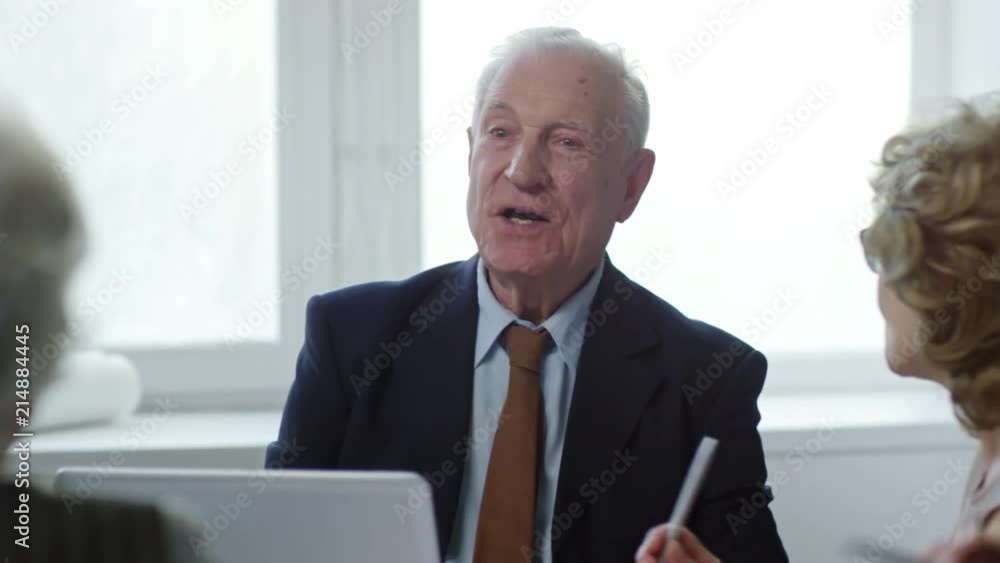PAN with slow motion of elderly businessman in suit and necktie talking on meeting with senior colleagues in office PAN with slow motion of elderly businessman in suit and necktie talking on meeting with senior colleagues in office