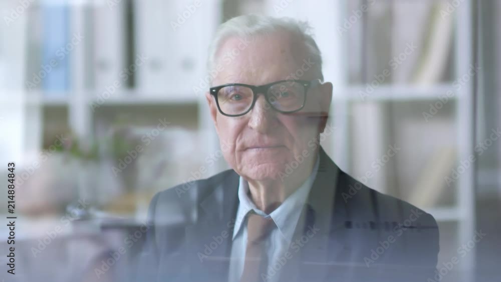 Tilt up shot through glass window in office: senior business executive with grey hair sitting at his desk and looking t camera Tilt up shot through glass window in office: senior business executive with grey hair sitting at his desk and looking t camera