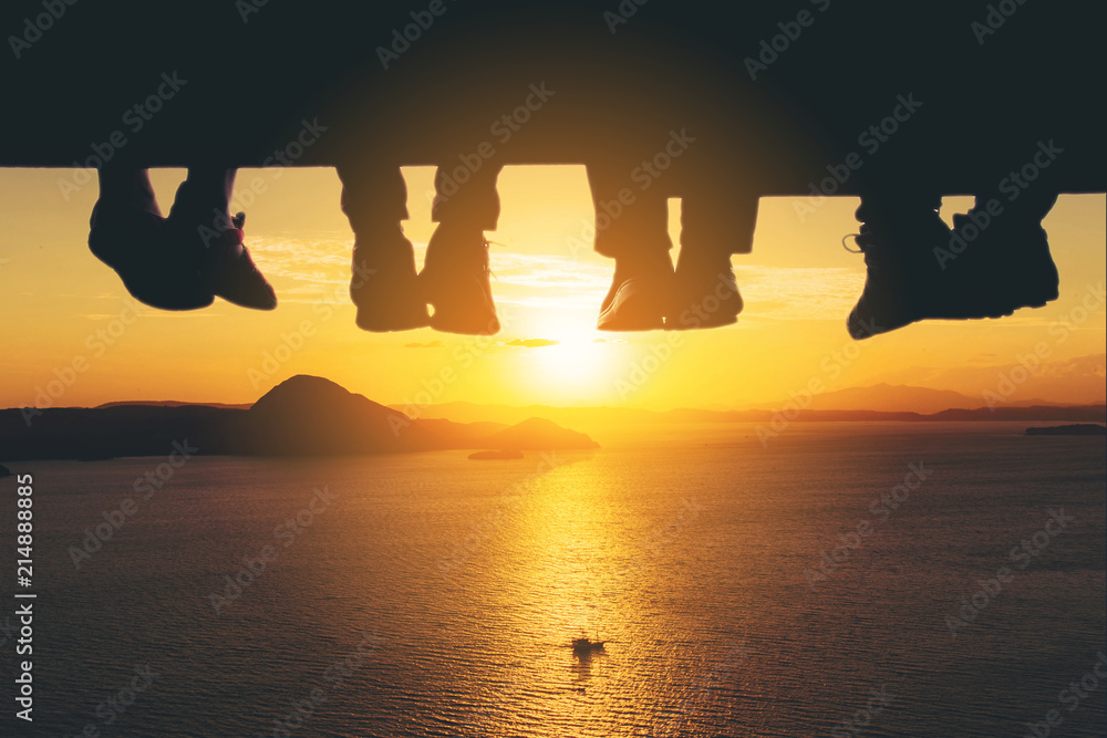 Φωτογραφία Underside view of dangling Four people legs with wearing trekking shoes sitting on the edge of wooden board-walk against a background of sea and sunset