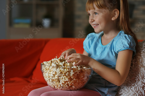 Little girl eating popcorn while watching TV on sofa in evening