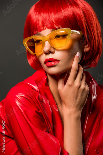 close-up portrait of fashionable young woman in red latex jacket and vintage yellow sunglasses isolated on grey