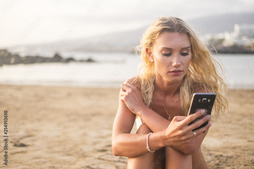 beautiful blonde young woman at the beach under the sunlight relaxed and connect the phone to internet to write and read messages with friends at home. travel people summer holiday concept beautiful blonde young woman at the beach under the sunlight relaxed and connect the phone to internet to write and read messages with friends at home. travel people summer holiday concept