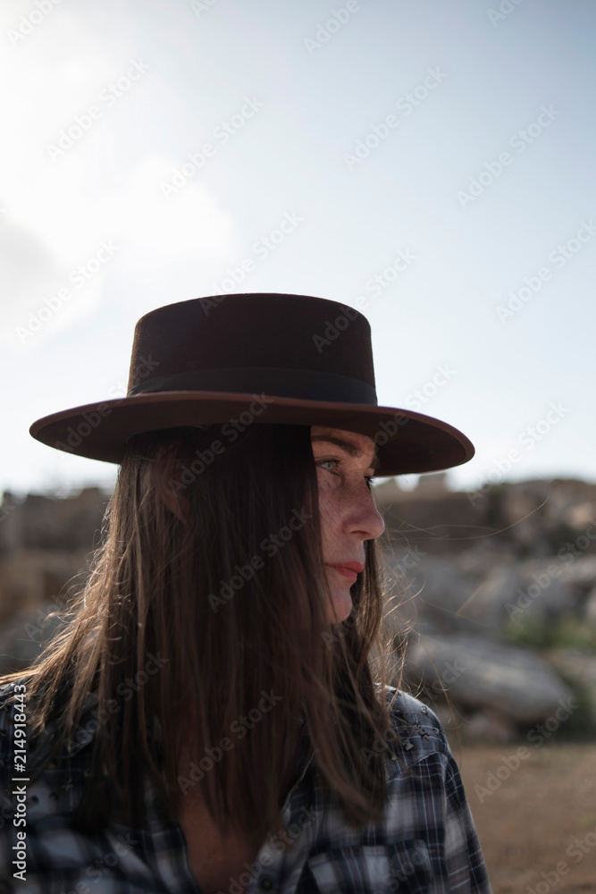 Brunette woman in brown hat and blue checkered shirt outdoor in sunlight.