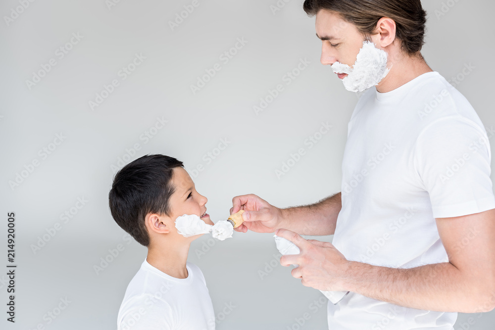 side view of father with brush putting shaving foam on sons face on grey background