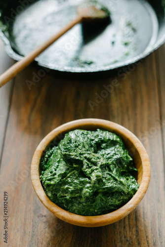 Portion of baby spinach as a side dish in a wooden bowl on a wooden background beside empty cast-iron pan.