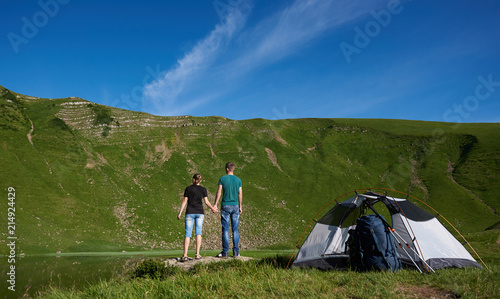Back view of a man and a woman stand near camping with a tent, backpacks and trekking sticks, looking at a mighty green mountain at the foot of which is a lake under a blue sky