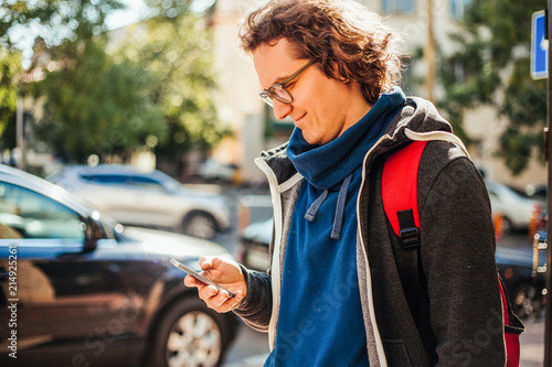 Young male tourist in eyeglasses standing on parking and using smartphone. Blurred cars background