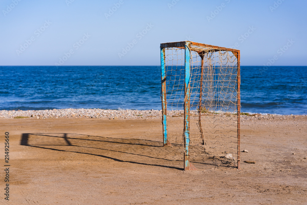 Old vacant football soccer goal gate on front of the blue ocean. Old sports field with rusty goal and net on meadow with muddy front line.