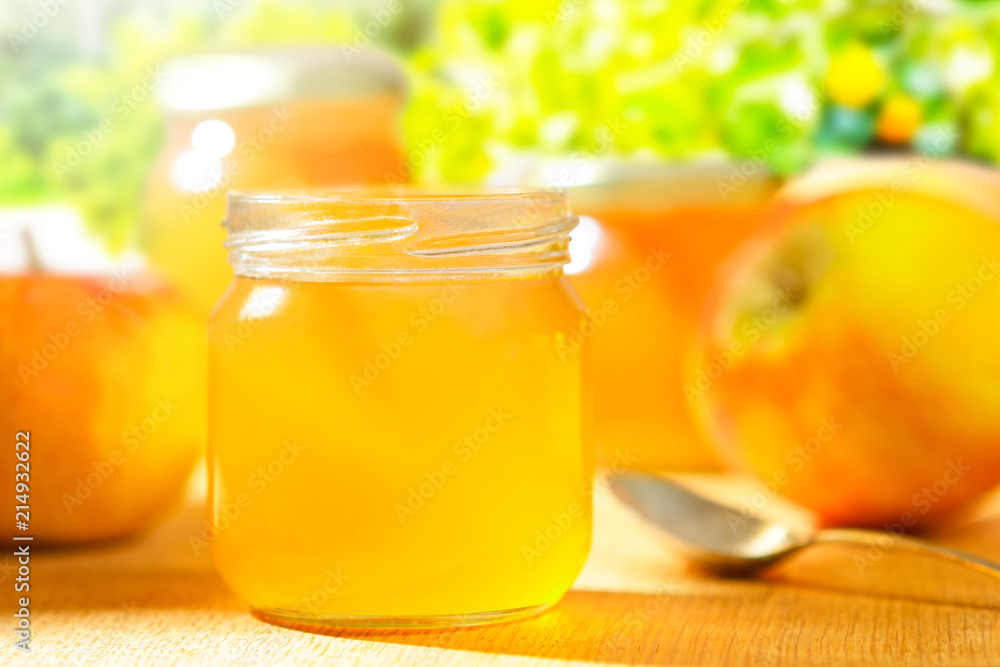 Homemade apple jelly in glass jars with fresh apples and a spoon on a wooden table in front of trees in bright sunshine Homemade apple jelly in glass jars with fresh apples and a spoon on a wooden table in front of trees in bright sunshine