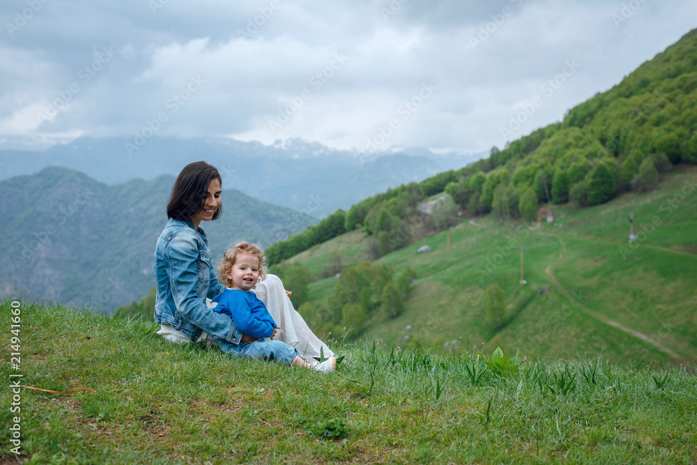 Young mother and her little baby daughter sitting on the green grass on meadow and enjoying beautiful mountains. Sky with clouds
