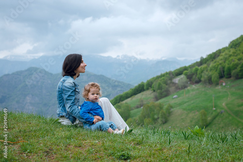 Young brunette mother and her little baby daughter sitting on the green grass on meadow and enjoying beautiful mountains. Sky with clouds
