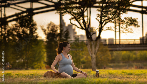 Beautiful pregnant young woman with little dog sitting on mat, enjoying yoga, relaxing, feeling alive, breathing fresh air, calm and dreaming, in green summer park. on the background of the railway