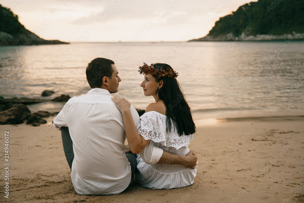loving couple in white sit on the beach on a sandy beach in the evening and look at each other. A tropical holiday for two.