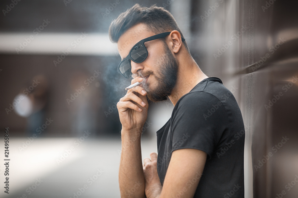 Fototapeta premium A young handsome white bearded man in sunglasses and black t-shirt smokes a cigarette in the street in the spring. Close up.
