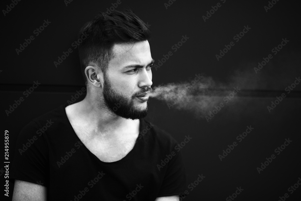A young handsome white bearded man in black t-shirt smokes a cigarette in the street in the spring. Close up. Black and white.