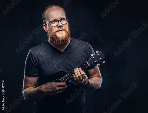 Redhead bearded male musician wearing glasses dressed in a gray t-shirt playing on a ukulele. Isolated on a dark textured background.