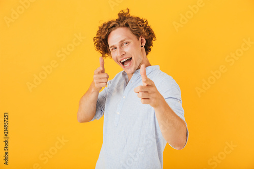 Photo of amusing handsome man 20s smiling and pointing fingers at you, isolated over yellow background
