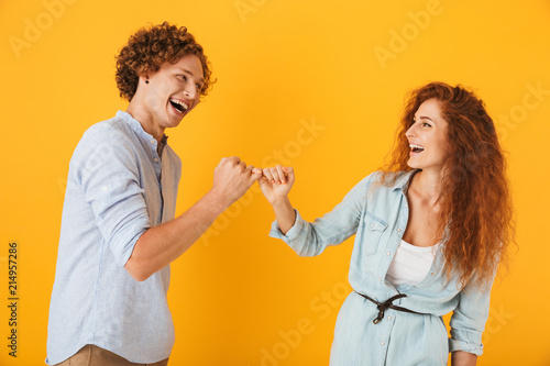 Photo of happy people man and woman smiling and hook each other's little fingers in conciliation or friendship, isolated over yellow background