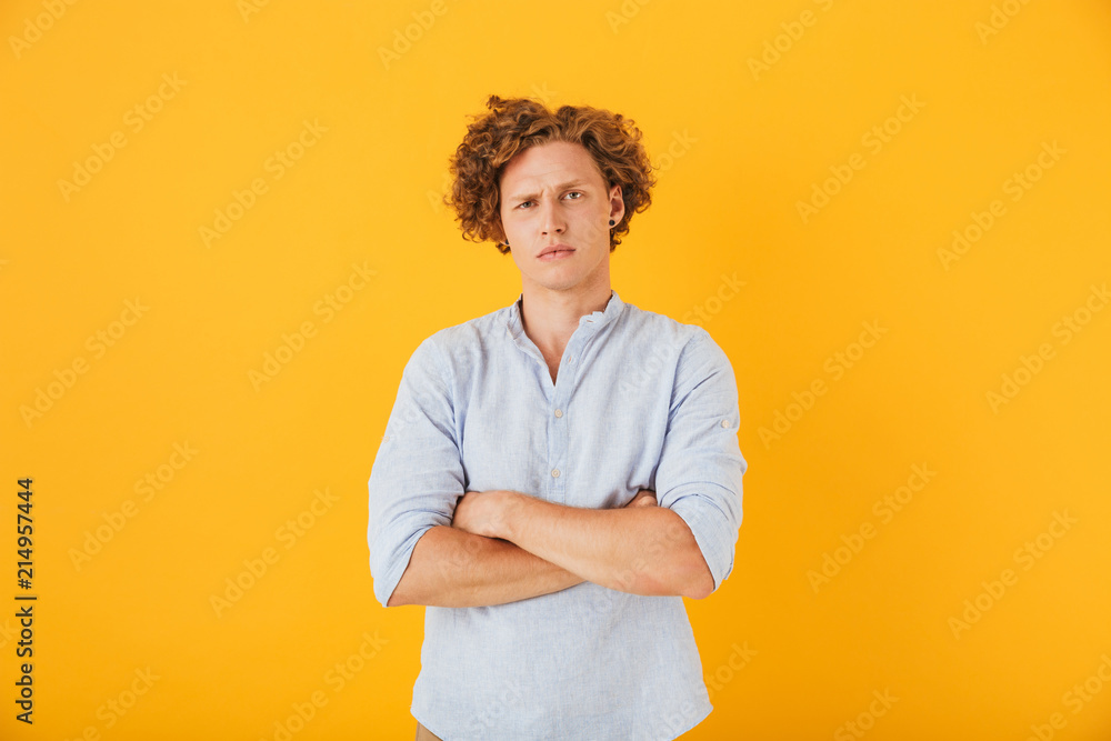 Photo of upset serious man 20s with curly hair standing with arms folded, isolated over yellow background Photo of upset serious man 20s with curly hair standing with arms folded, isolated over yellow background