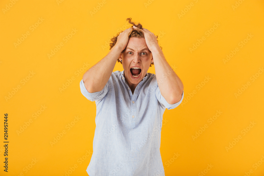 Photo of shocked young man 20s with curly hair grabbing head and shouting in anger, isolated over yellow background Photo of shocked young man 20s with curly hair grabbing head and shouting in anger, isolated over yellow background