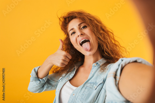 Image of funny joyous woman 20s showing thumb up with tongue sticking out while taking selfie photo, isolated over yellow background © Drobot Dean