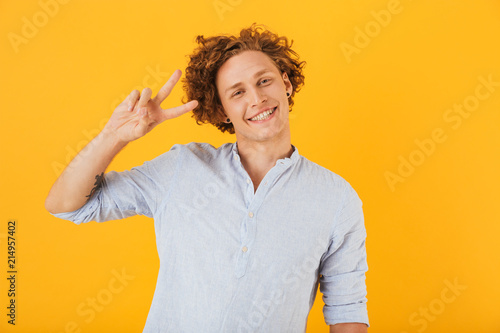 Image of young cheerful man 20s smiling and showing peace sign at camera, isolated over yellow background