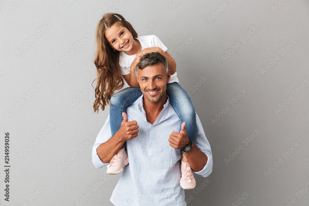 Portrait of adorable family, daughter smiling and sitting on the neck of her father 30s, isolated over gray background - obrazy, fototapety, plakaty 