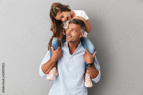 Portrait of lovely daughter smiling and sitting on the neck of her handsome father, isolated over gray background
