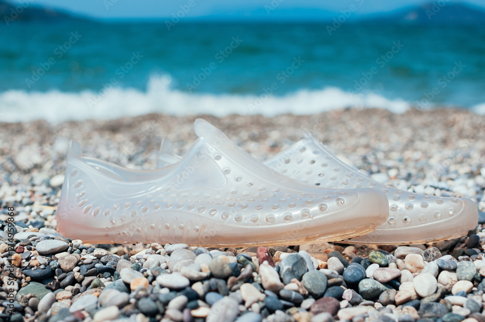 Pair of transparent swimming shoes on marble pebble beach, turquoise sea  water in the background. Summer holiday concept. Kids beach shoes. Soft,  lightweight material aqua shoes. Dotted water shoes Stock Photo