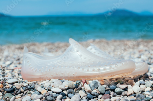 Pair of transparent swimming shoes on marble pebble beach, turquoise sea water in the background. Summer holiday concept. Kids beach shoes. Soft, lightweight material aqua shoes. Dotted water shoes
