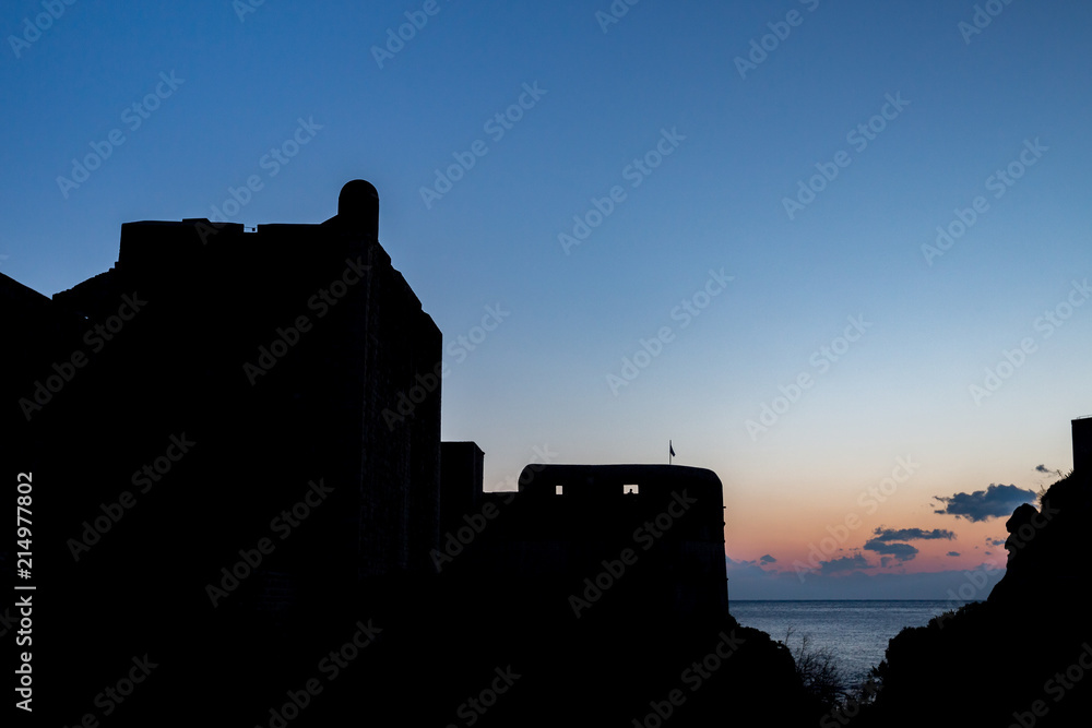 Obraz premium The black silhouette of the Dubrovnik castle tower on sunset with the colorful sky and the dark blue Adriatic Sea, almost clear sky with few small clouds