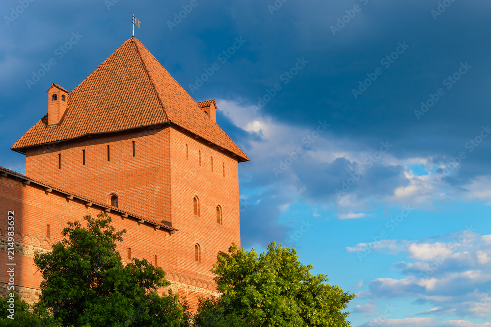 A powerful tower of the Lida castle from red brick and stone lit with  the sun against a beautiful but stormy sky