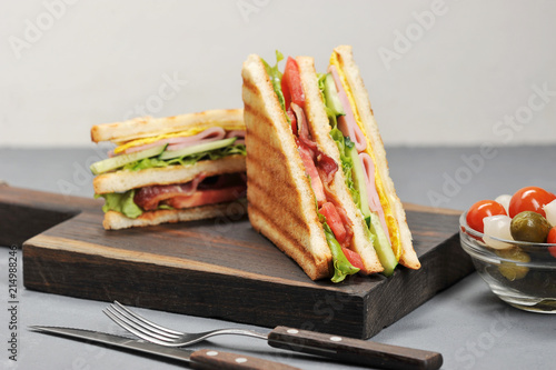 Classic club sandwich with ham and bacon on a wooden board. Next cup is a mixture of pickled miniature onions, gherkins, tomatoes. Gray background. Close-up. Macro photography.