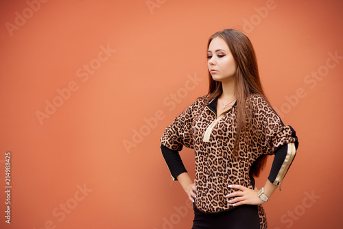 The brown-haired woman in fashionable leopard clothes is holding her hands on her belt against the red wall. Female portrait in fashion style on the street.